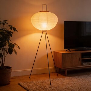 May include: A modern floor lamp with a paper lantern shade. The lamp has a round, cream-colored shade supported by a black tripod base. The lamp is in a living room setting with a wooden cabinet and a potted plant.