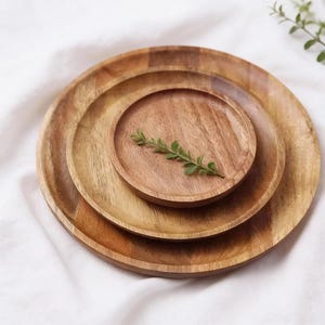May include: Three wooden plates of different sizes, nested together. The plates are round with a natural wood grain finish. A small sprig of greenery rests on the smallest plate. The plates are arranged on a white surface.