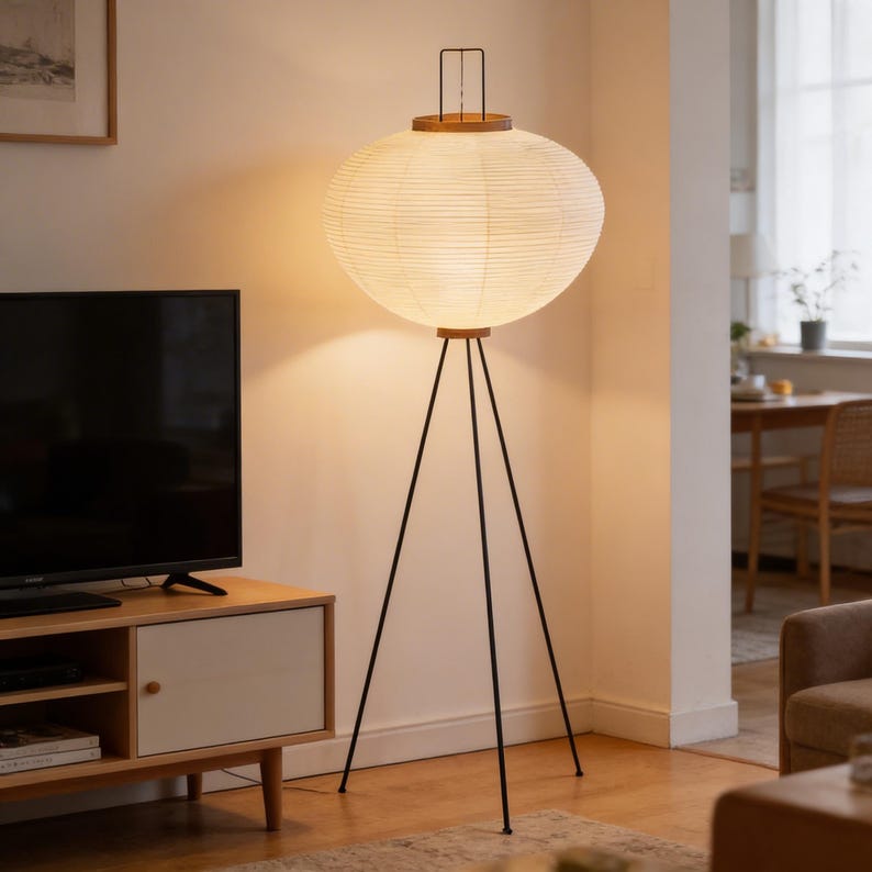 May include: A modern floor lamp with a large, round, paper lantern shade. The shade is cream-colored and rests on a wooden base and black metal tripod legs. The lamp is a stylish addition to any living space.