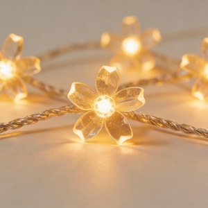 以下が含まれることがあります： 暖白色LEDフェアリーライトのストリングのクローズアップ。各ライトは、明るい中心部を持つ半透明の花の形をしています。ワイヤーはねじれたシルバー色で、背景は柔らかくニュートラルな色調です。