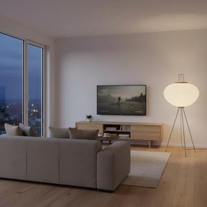 May include: A modern living room with a light gray sofa, a wooden entertainment center, and a large window. A floor lamp with a white, oval shade and a dark metal tripod base provides soft lighting. A TV displays a landscape scene.
