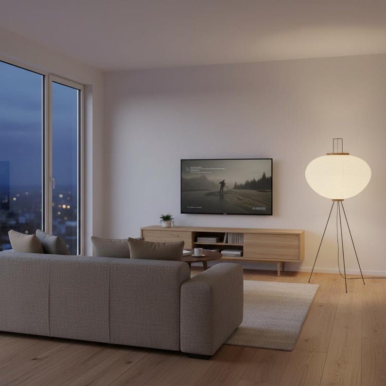 May include: A modern living room with a light gray sofa, a wooden entertainment center, and a large window. A floor lamp with a white, oval shade and a dark metal tripod base provides soft lighting. A TV displays a landscape scene.