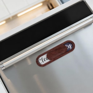 Könnte beinhalten: Ein hölzerner Geschirrspülmagnet mit einem Schiebedeckel. Der Deckel ist aus dunklem Holz, und die Wörter "Clean" und "Dirty" sind sichtbar. Der Magnet ist an einem Edelstahlgerät befestigt.