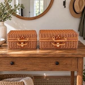 May include: Two rectangular woven wicker suitcases with light brown finish. Each case features a hinged lid, a matching woven handle, and a central tan leather-like latch mechanism with silver-toned metal hardware and rivets.