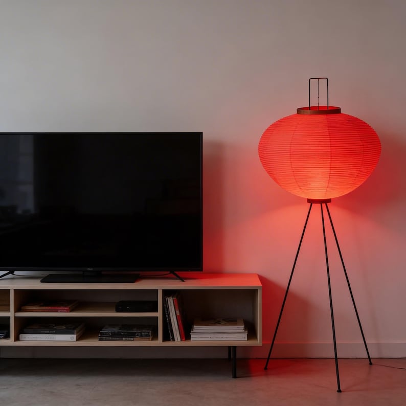 May include: A red paper lantern floor lamp with a black tripod base. The lamp has a round shape and emits a warm glow. The lamp is next to a light wood TV stand and a large black television.