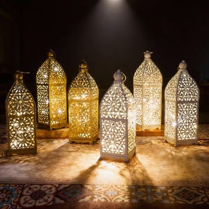 May include: Several decorative metal lanterns with detailed, cut-out designs. The lanterns are in gold and white, lit from inside, producing a warm light. They are arranged on a patterned surface, ideal for home decoration. The lanterns are approximately 30 cm tall.