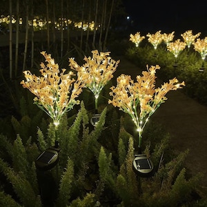 May include: Solar-powered garden lights in the shape of yellow flowers with small white lights. The lights are placed in a green bush. The lights are illuminated at night. Each light has a black solar panel to absorb sunlight.