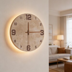 May include: A round wall clock with a beige stone-like face and brown numerals. The clock hands are brown, and the clock is illuminated with a warm light around the back. The clock is mounted on a white wall.