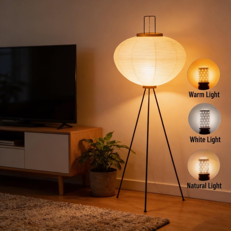 May include: A modern floor lamp with a paper lantern shade and a black tripod base. The lamp has a warm, white, and natural light setting. The lamp is in a living room setting with a TV and a plant.