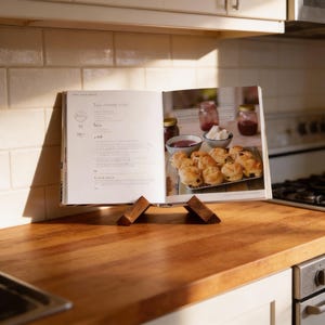 May include: A wooden cookbook stand supports an open cookbook on a wooden kitchen surface. The cookbook shows a recipe with text and a photo of baked goods, jam, and cream. The stand is made of two triangular wooden pieces.