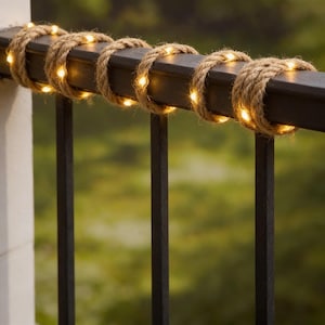 May include: A black metal railing section adorned with natural rope and small, warm white LED lights. The rope is wrapped around the railing, with the lights providing a gentle illumination. The background shows blurred green foliage.