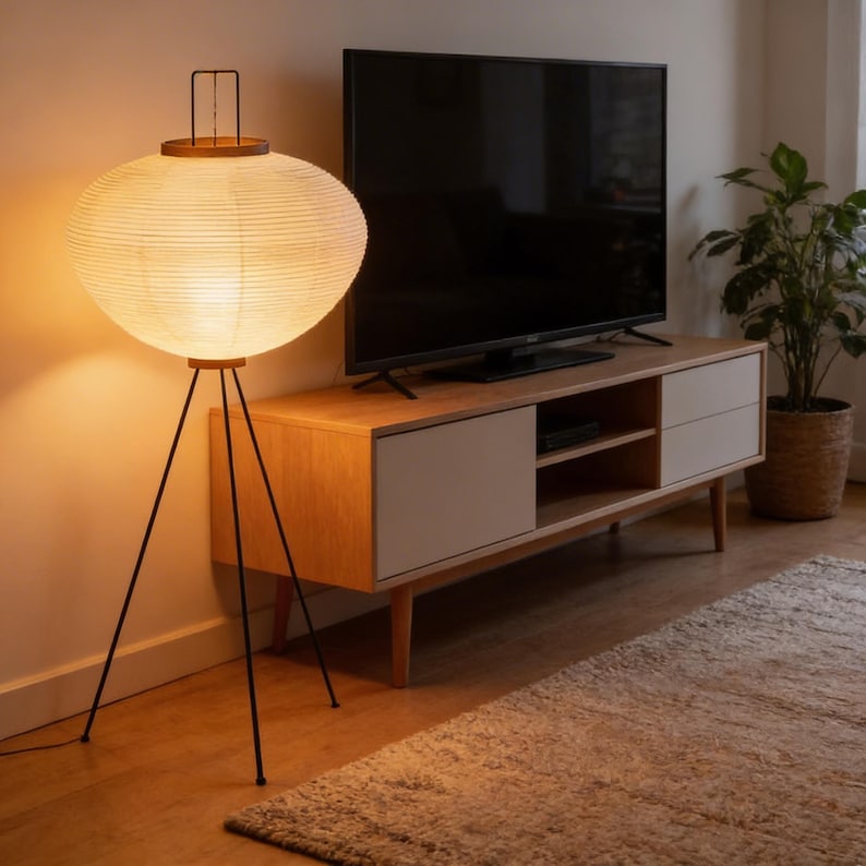 May include: A modern floor lamp with a large, round, cream-colored paper shade. The lamp has a dark metal tripod base and a wooden top. The lamp is next to a wooden TV stand with a black TV on top.