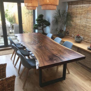 May include: A large, live-edge wooden dining table with a dark brown finish. The table is supported by a black metal base and surrounded by gray upholstered chairs. The table measures approximately 100 inches long.