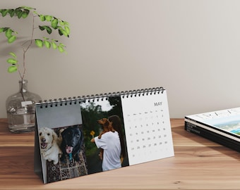 May include: A desk calendar featuring photos of dogs, with the month of May displayed. The calendar is spiral-bound and stands on a wooden surface. A clear glass vase with a plant is in the background.