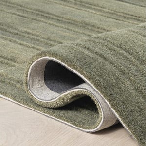 Könnte beinhalten: Ein aufgerollter, olivgrüner Teppich mit einem strukturierten, gerippten Design. Die Unterseite des Teppichs ist ein grauer Stoff mit weißem Rand. Der Teppich liegt auf einer hellen Holzoberfläche.