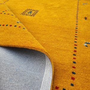 May include: A mustard yellow area rug with a soft, textured pile surface. The rug features small, scattered geometric patterns in red and blue, including a central square motif and a vertical border line. The underside shows a light grey backing.