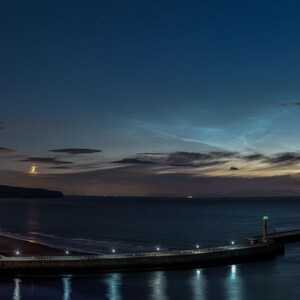 Puede incluir: Paisaje costero nocturno con cielo azul oscuro y nubes. La imagen muestra un pueblo con edificios iluminados, un muelle con luces y un mar en calma que refleja las luces. El cielo tiene colores de atardecer.