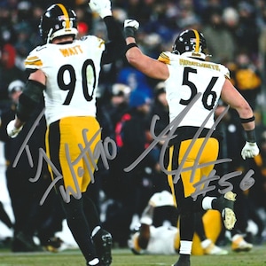 May include: Two football players in black and gold uniforms celebrate on a green field. The player on the left wears the number 90, and the player on the right wears the number 56. Both players have their arms raised in the air.