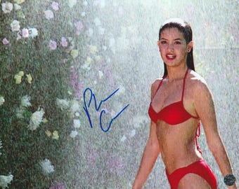 May include: A glossy photograph featuring a person with dark hair wearing a bright red two-piece bikini. The person is positioned in front of a blurred background of green foliage and white flowers under a light mist, with a blue ink signature overlay.
