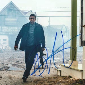 May include: A photograph of a person wearing a dark jacket and grey shirt, holding a black rifle, with a blue ink signature across the image. The background shows a building with the text Dennis Crowley Marine Supplies and Services visible.