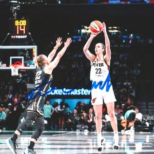 May include: A basketball player in a white and blue uniform with the number 22 shoots a basketball. Another player in a black and gold uniform attempts to block the shot. The background shows a basketball court and spectators.