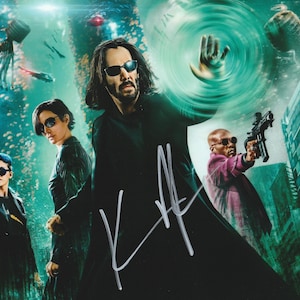 May include: A promotional poster for The Matrix Resurrections featuring Keanu Reeves in a black coat with a silver autograph, alongside Carrie-Anne Moss, Jessica Henwick, and Yahya Abdul-Mateen II, all wearing sunglasses against a green digital background.