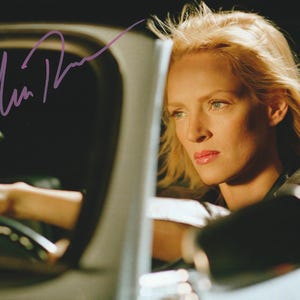 May include: A glossy photograph featuring a blonde individual with a serious expression, seated in the driver's seat of a vehicle. The image includes a purple ink signature across the upper left portion and a small circular emblem in the bottom corner.