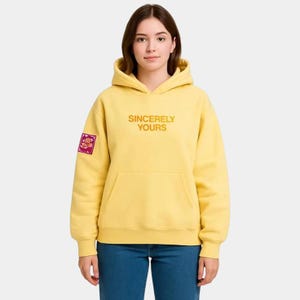 Op de afbeelding: Een gele hoodie met de tekst "Sincerely Yours" in oranje. De hoodie heeft een kangoeroezak en een kleine patch op de linkermouw. De persoon draagt een blauwe spijkerbroek.