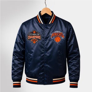 Pode incluir: Uma jaqueta universitária de cetim azul-marinho com fecho de botões de pressão, gola, punhos e bainha canelados às riscas. O peito apresenta um emblema 20 NBA 26 Champions à direita e o logótipo dos New York Knicks à esquerda.