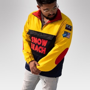 Puede incluir: Chaqueta anorak amarilla, negra y azul marino con detalles en rojo. La chaqueta presenta el texto "SNOW BEACH" en rojo en la parte delantera, junto con "POLO SPORT" y un parche de la bandera estadounidense. La chaqueta tiene cierre de media cremallera y cuello alto.