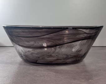 May include: A decorative glass bowl with a smoky gray swirl pattern. The bowl has a wide, shallow shape and is made of transparent glass. The gray pattern creates a marbled effect, adding visual interest to the piece. The bowl is suitable for serving or display.