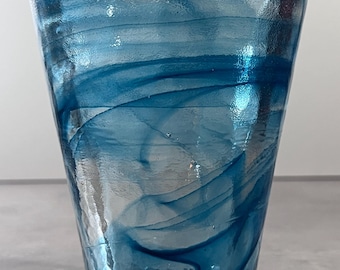Op de afbeelding: Een heldere glazen tumbler met een uitlopende bovenkant en een getextureerd oppervlak. Wervelingen van blauw en blauwgroen glas zijn zichtbaar in het hele glas. De bodem van het glas heeft een moeilijk leesbare tekst.