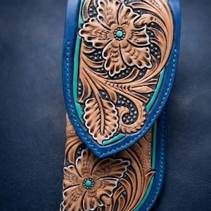 May include: Blue leather knife sheath with floral tooling. The sheath features a turquoise accent and a detailed carved floral design. The edges are blue, and the background is a dark blue.
