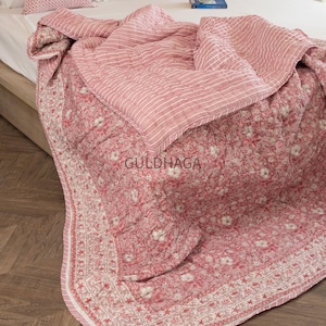 Op de afbeelding: Een omkeerbare, doorgestikte sprei met een roze en witte bloemenprint aan de ene kant en een roze en wit gestreept patroon aan de andere kant. De stof heeft een zichtbare stiktextuur. Het woord GULDHAGA staat in het midden afgedrukt.