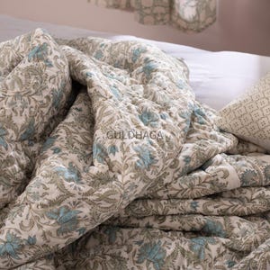 May include: A quilted bedspread with a floral pattern in shades of blue and green on a cream background. The bedspread is folded and draped over a bed, with the text "GULDHAGA" visible on the fabric.