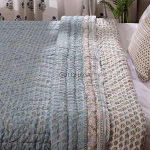 Könnte beinhalten: Eine hellblaue und cremefarbene Steppdecke mit floralem Muster und dem Text "GULDHAGA". Die Steppdecke ist über einem Bett gefaltet und hat einen detaillierten Rand. Das Kissen hat ein passendes Blumenmuster.