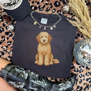 Puede incluir: Una camiseta gris carb&oacute;n oscuro con un gr&aacute;fico bordado centrado de un perro doodle de color marr&oacute;n dorado sentado. La camiseta se muestra con un collar de cadena dorado, una gorra de b&eacute;isbol negra y una bota de cuero negra con bordado floral blanco.