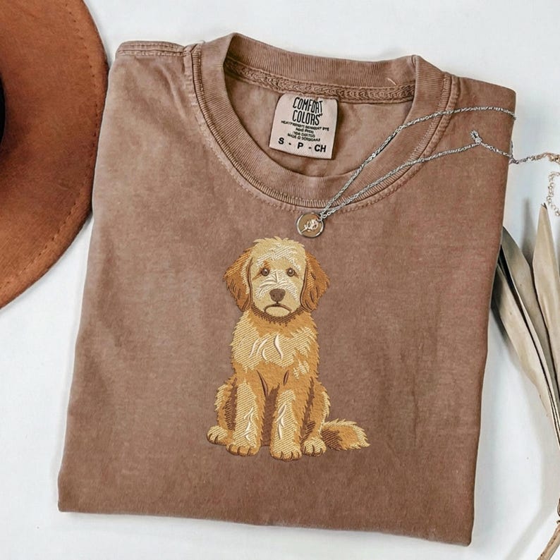 Puede incluir: Una camiseta de manga corta marr&oacute;n con un perro de color dorado bordado sentado en el centro. La etiqueta interior del cuello dice Comfort Colors, S-P-CH. Un collar de cadena plateada fina con un peque&ntilde;o colgante circular descansa en el cuello.