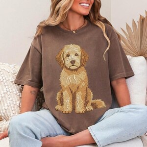 Puede incluir: Una camiseta marr&oacute;n de manga corta con una ilustraci&oacute;n gr&aacute;fica centrada de un perro de color dorado con pelaje texturizado. El perro est&aacute; representado en posici&oacute;n sentada, mirando hacia adelante con una expresi&oacute;n neutral. La camiseta tiene cuello redondo.