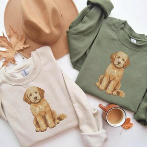 Puede incluir: Una sudadera beige de cuello redondo y una sudadera verde oliva de cuello redondo, cada una con un perro golden doodle bordado. Un sombrero beige de ala ancha, una hoja de arce seca y una taza de cer&aacute;mica con caf&eacute; acompa&ntilde;an a las prendas.
