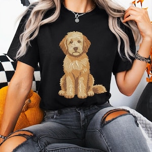Puede incluir: Una camiseta negra de manga corta con un bordado central detallado de un perro de color dorado sentado. El perro tiene pelaje texturizado, orejas ca&iacute;das y una nariz marr&oacute;n. La camiseta se combina con unos vaqueros grises desgastados.