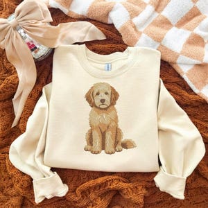 Puede incluir: Una sudadera de cuello redondo color crema con un bordado detallado y centrado de un perro doodle de color marr&oacute;n dorado sentado. El perro tiene pelaje desgre&ntilde;ado, orejas ca&iacute;das y una expresi&oacute;n dulce. La sudadera tiene mangas largas.