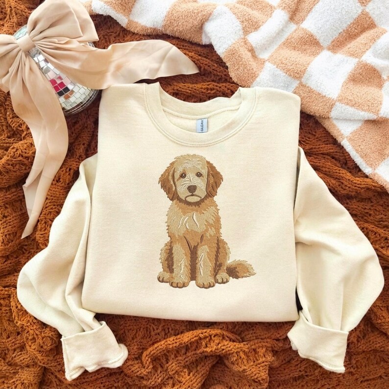 Puede incluir: Una sudadera de cuello redondo color crema con un bordado detallado y centrado de un perro doodle de color marr&oacute;n dorado sentado. El perro tiene pelaje desgre&ntilde;ado, orejas ca&iacute;das y una expresi&oacute;n dulce. La sudadera tiene mangas largas.