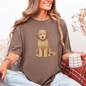 Puede incluir: Una camiseta marr&oacute;n de manga corta con una ilustraci&oacute;n gr&aacute;fica centrada de un perro de color dorado sentado. El perro tiene un aspecto texturizado y peludo con tonos de pelaje beige claro y marr&oacute;n dorado, representado de frente.