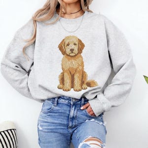 Puede incluir: Una sudadera gris claro de cuello redondo con un bordado detallado y centrado de un perro doodle de color marr&oacute;n dorado sentado. El perro tiene pelaje texturizado, orejas ca&iacute;das y una expresi&oacute;n neutra. Se combina con pantalones vaqueros azules.