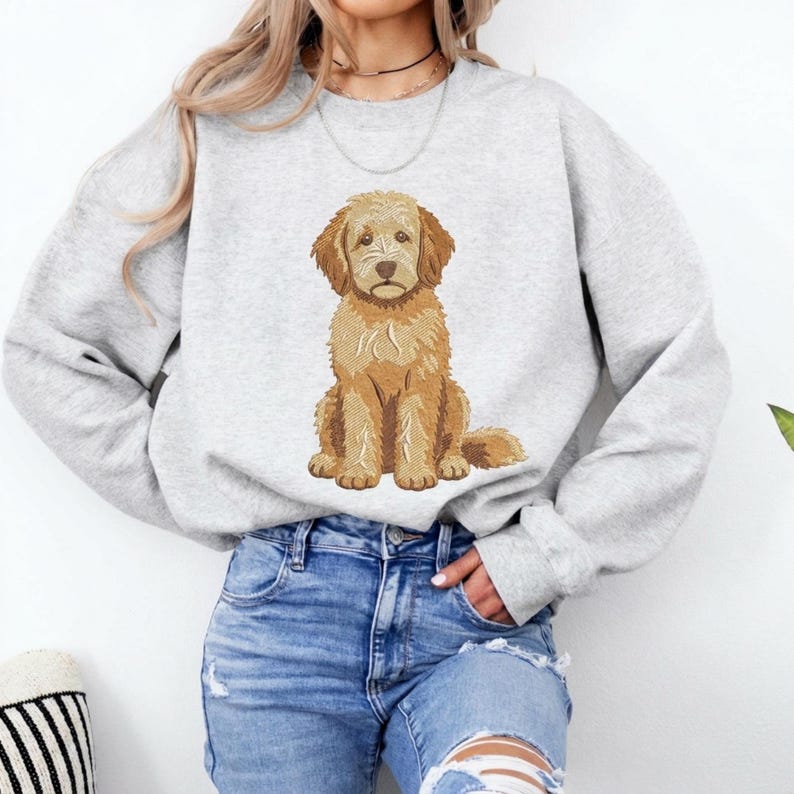 Puede incluir: Una sudadera gris claro de cuello redondo con un bordado detallado y centrado de un perro doodle de color marr&oacute;n dorado sentado. El perro tiene pelaje texturizado, orejas ca&iacute;das y una expresi&oacute;n neutra. Se combina con pantalones vaqueros azules.