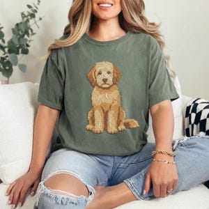 Puede incluir: Una camiseta de manga corta color verde salvia con una ilustraci&oacute;n gr&aacute;fica centrada de un perro doodle de color marr&oacute;n dorado sentado. El perro tiene pelaje rizado y texturizado, orejas ca&iacute;das y una nariz oscura, en un estilo detallado.