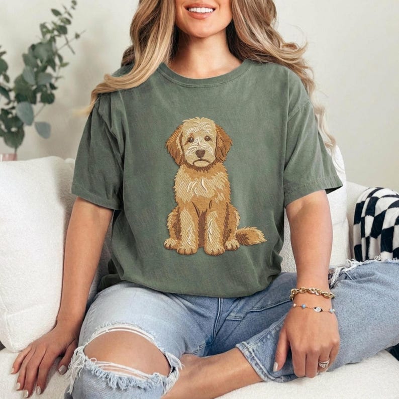 Puede incluir: Una camiseta de manga corta color verde salvia con una ilustraci&oacute;n gr&aacute;fica centrada de un perro doodle de color marr&oacute;n dorado sentado. El perro tiene pelaje rizado y texturizado, orejas ca&iacute;das y una nariz oscura, en un estilo detallado.