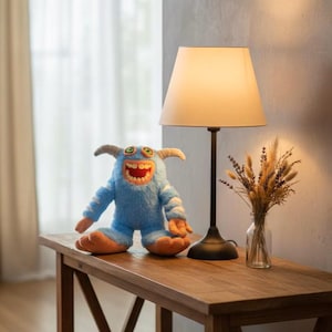 Peut inclure: Un jouet monstre en peluche bleu avec des pieds orange et un large sourire édenté est posé sur une table en bois. Une lampe noire avec un abat-jour beige et un vase de fleurs séchées sont également sur la table.