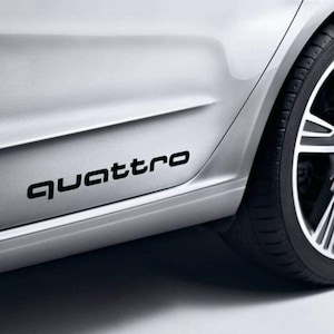 May include: Black "Quattro" decal applied to the side of a silver car. The decal is positioned near the bottom of the car door, with a portion of the tire visible on the right side.